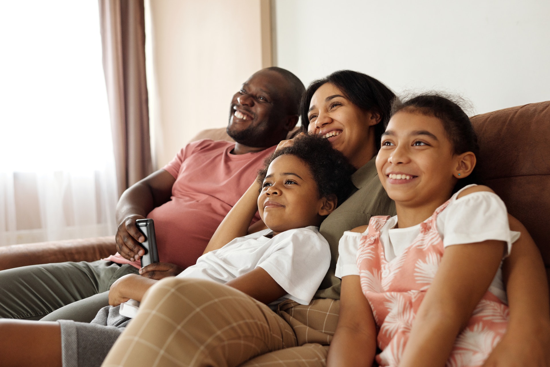 A smiling family of four sits closely together on a couch, watching TV. The father holds a remote, and the mother, son, and daughter all look happy and relaxed in a brightly lit living room.